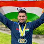 An image of Anish Bhanwala wearing 3 medals and holding the Indian Flag above his head